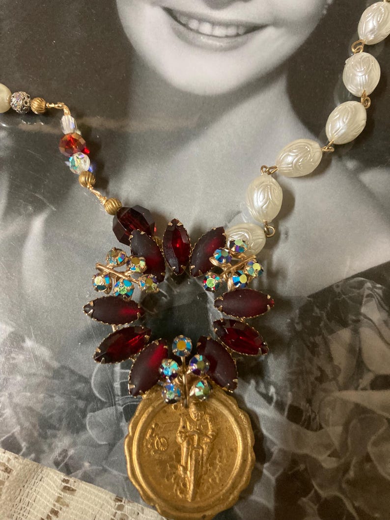 Op de afbeelding: Vintage ketting met een gouden hanger met een figuur, omringd door donkerrode en iriserende kristallen. De ketting bevat witte, getextureerde kralen en kleinere, kleurrijke kralen. Het ontwerp is geplaatst tegen een vintage foto.