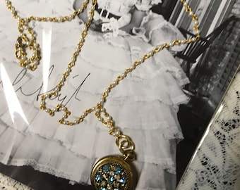 May include: A gold chain necklace with a round pendant featuring blue and clear rhinestones. The necklace is on top of a black and white photo of a young woman wearing a white dress.