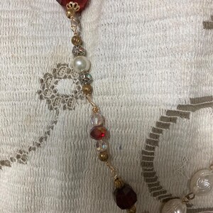 Op de afbeelding: Een goudkleurige ketting met een verscheidenheid aan kralen en edelstenen. De ketting bevat een grote, facetgeslepen, rode edelsteen, parels en kleinere, iriserende kralen. Het ontwerp bevat goudkleurige metalen accenten en een bloemvormige hanger met rode en iriserende stenen.