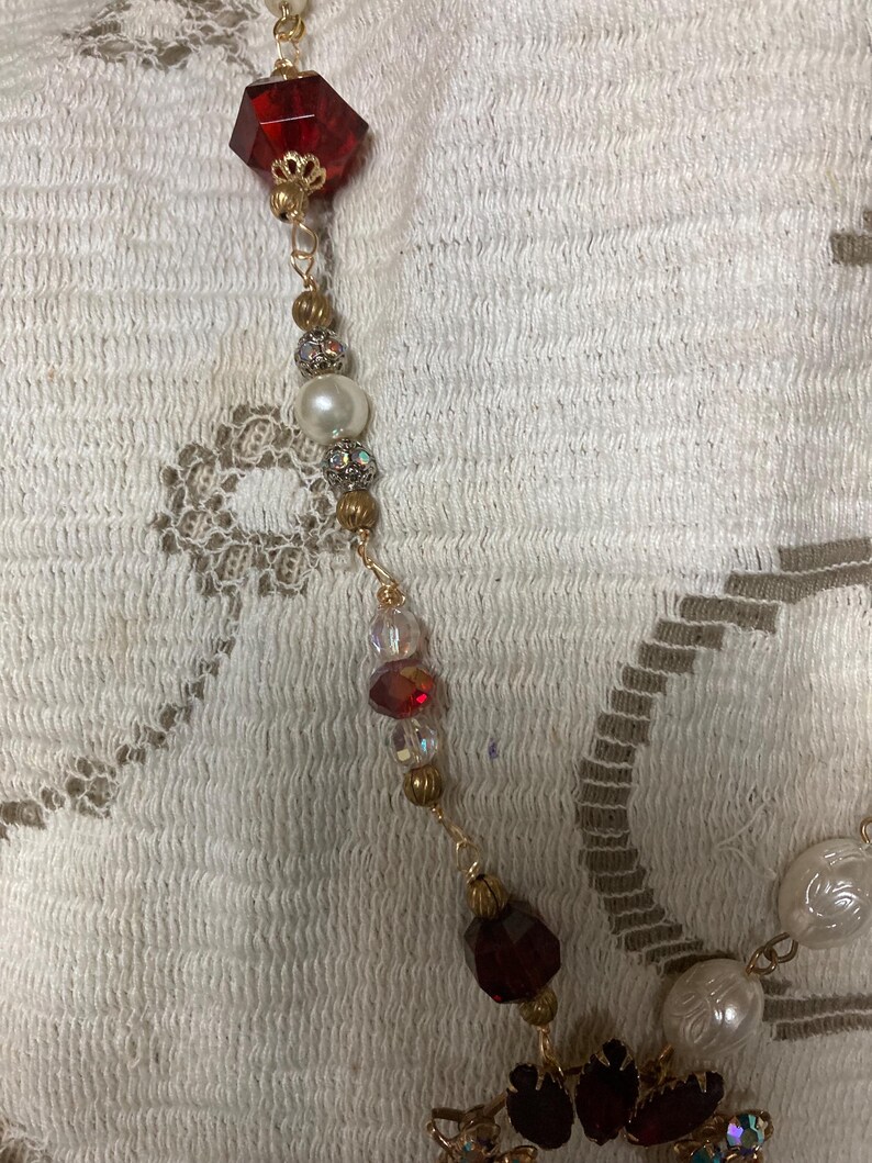 Op de afbeelding: Een goudkleurige ketting met een verscheidenheid aan kralen en edelstenen. De ketting bevat een grote, facetgeslepen, rode edelsteen, parels en kleinere, iriserende kralen. Het ontwerp bevat goudkleurige metalen accenten en een bloemvormige hanger met rode en iriserende stenen.