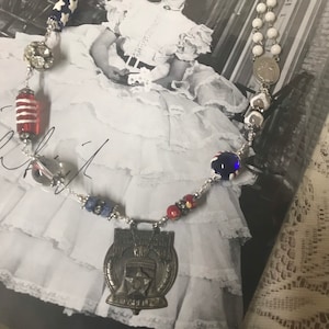 May include: A silver necklace with a variety of beads, including red, white, and blue beads, a pearl, and a silver charm with a star and the text "The American Legion 1919".