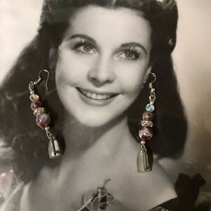 May include: A pair of silver earrings with a red and white marbled bead, a silver bead with a clear rhinestone, and a silver charm shaped like a boat. The earrings are hanging from a black and white photo of a woman.