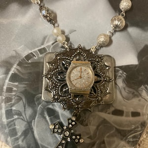 May include: A silver-toned necklace featuring a watch pendant with a decorative frame and a cross pendant. The necklace has a beaded chain with various silver and pearl-like beads. The watch has a silver face and gold hands.