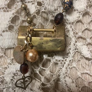 May include: A vintage-style necklace featuring a brass-toned rectangular pendant with a small heart charm. The necklace includes a mix of gold-toned beads, faceted dark brown beads, and a faux pearl. The background is a cream-colored lace.
