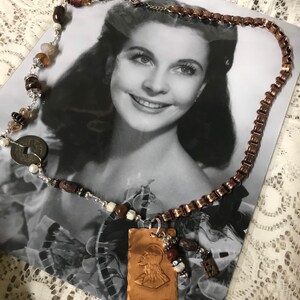 May include: A copper chain necklace with a copper pendant featuring a profile of a person with long hair. The pendant is attached to a brown leather cord with brown and white beads. The necklace is lying on a black and white photograph of a woman with dark hair. The photograph is signed "Vivien Leigh".