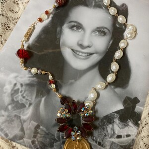 Op de afbeelding: Een vintage-stijl ketting met rode, witte en gouden kralen, met een centrale hanger van een gouden munt en een cluster van rode en iriserende stenen. De ketting wordt getoond op een zwart-wit foto.