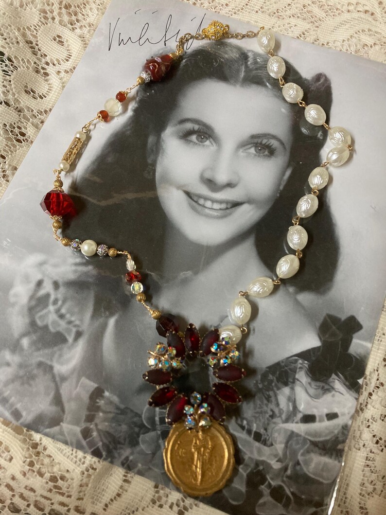 Op de afbeelding: Een vintage-stijl ketting met rode, witte en gouden kralen, met een centrale hanger van een gouden munt en een cluster van rode en iriserende stenen. De ketting wordt getoond op een zwart-wit foto.