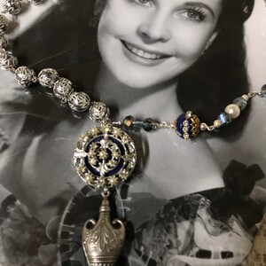 May include: A silver necklace with a blue and silver pendant shaped like a bottle. The pendant is adorned with small white pearls and a blue enamel design. The necklace is made of silver beads and a silver chain.