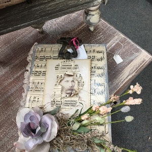 May include: A vintage-style wooden box decorated with sheet music and a floral arrangement. The box features a torn photo of a man with a mustache and a small tag attached to the side.