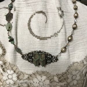 May include: A necklace with a silver chain and green glass beads. The necklace has a pendant in the shape of two small shoes. The pendant is made of metal and has a filigree design.