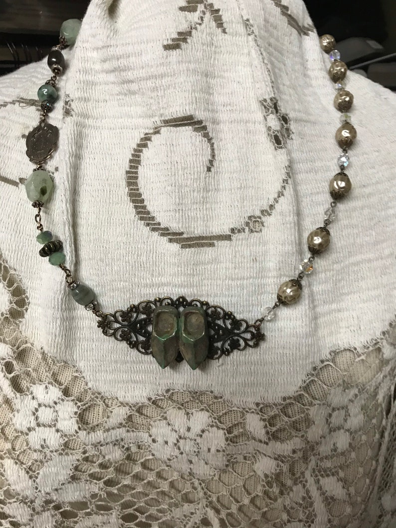 May include: A necklace with a silver chain and green glass beads. The necklace has a pendant in the shape of two small shoes. The pendant is made of metal and has a filigree design.