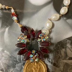 Op de afbeelding: Vintage ketting met een gouden hanger met een figuur, omringd door donkerrode en iriserende kristallen. De ketting bevat witte, getextureerde kralen en kleinere, kleurrijke kralen. Het ontwerp is geplaatst tegen een vintage foto.