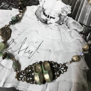 May include: A vintage-style necklace with green and brown beads, a metal filigree design, and two small silver shoes. The necklace is displayed on a black and white photo of a woman in a white dress.