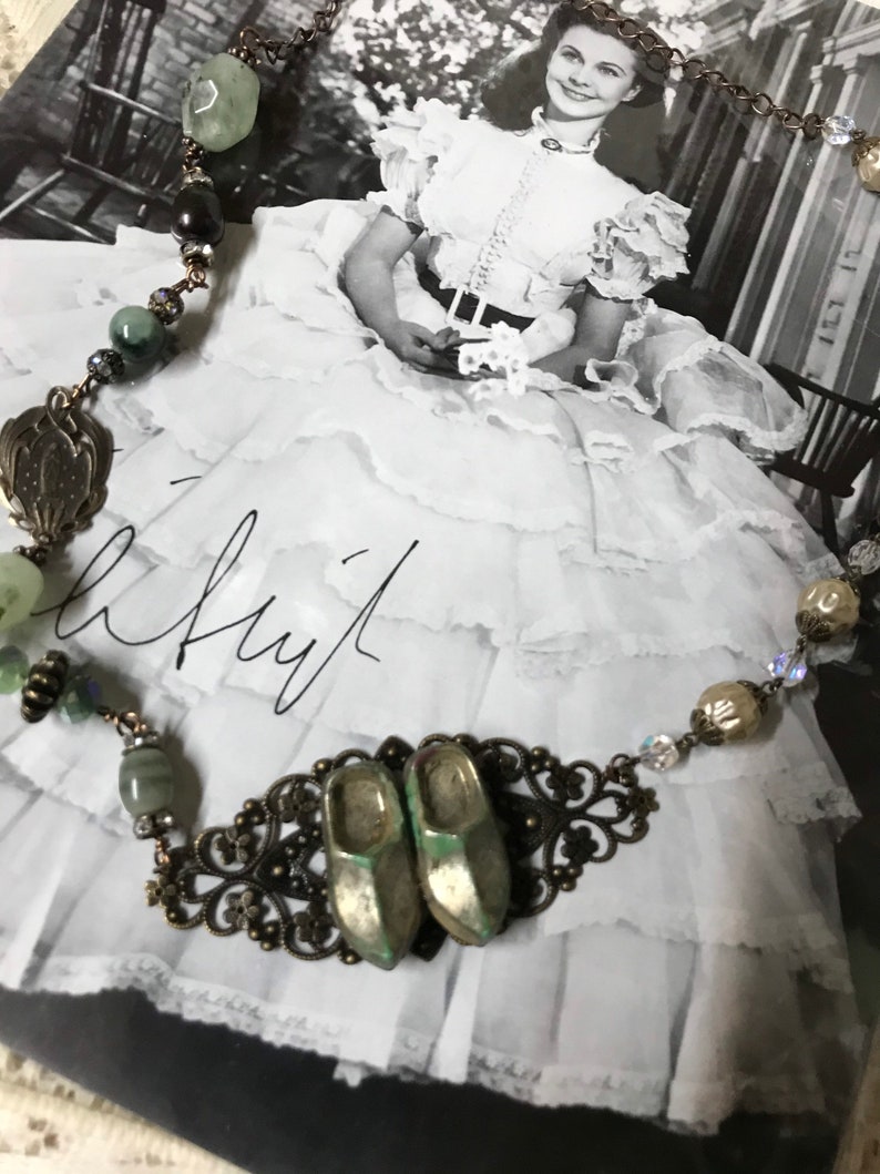 May include: A vintage-style necklace with green and brown beads, a metal filigree design, and two small silver shoes. The necklace is displayed on a black and white photo of a woman in a white dress.