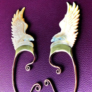 May include: A pair of gold and copper ear cuffs with a white and gold eagle design. The eagle is carved in detail and the cuffs are made of wire.