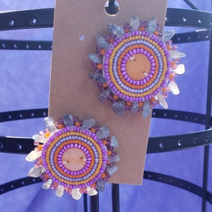 Puede incluir: Un par de pendientes de cuentas con un diseño circular. Los pendientes están hechos con cuentas moradas, naranjas y blancas y tienen un acento de piedra clara.