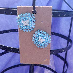 May include: A pair of blue and white beaded earrings with a white butterfly design. The earrings are displayed on a brown card.