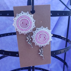 Può includere: Un paio di orecchini con perline rosa e bianche con un ciondolo a forma di fata argentata appeso a ciascun orecchino.