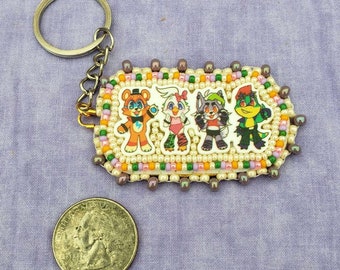 May include: A beaded keychain featuring four cartoon characters. The characters are a bear, a bird, a wolf, and a lizard. The keychain is oval shaped and has a silver key ring attached.