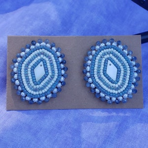 Può includere: Un paio di orecchini con perline blu e bianche con un pezzo centrale a forma di diamante bianco. Gli orecchini sono esposti su una carta marrone.