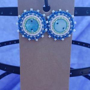 Può includere: Un paio di orecchini rotondi in perle blu, bianche e rosa con un centro verde e blu. Gli orecchini hanno un design a strati.