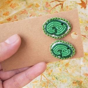 May include: A pair of green beaded earrings with a white center and a green border. The earrings are oval shaped and have a swirling pattern.