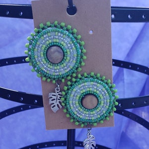 Puede incluir: Un par de pendientes de cuentas verdes y plateadas con un dije de hoja plateada que cuelga de cada pendiente.