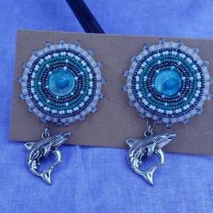 Puede incluir: Un par de pendientes de cuentas con un diseño circular azul y blanco y un dije de tiburón plateado que cuelga de cada pendiente.
