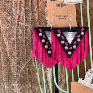May include: A pair of handmade earrings with a triangular design. The earrings feature a black top, a pink fringe, and silver-colored studs. The center has a patterned fabric. The earrings are displayed on a brown card with text.