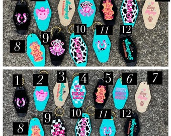 May include: A collection of novelty keychains in various colors and designs. The keychains feature images of a horseshoe, a smiling cowgirl, cow print, and text such as "Keys to the Shit Show" and "Somebody's Problem".