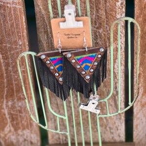 May include: A pair of triangular earrings with a brown leather top, colorful geometric design, and black fringe. The earrings have silver-colored studs and are displayed on a light green metal cactus-shaped stand.