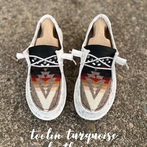 May include: Pair of slip-on shoes with a southwestern-inspired design. The shoes feature a white sole, a patterned fabric with a geometric design in shades of brown, red, and cream, and a black accent with decorative beading. The text "toolin turquoise leather co" is visible.
