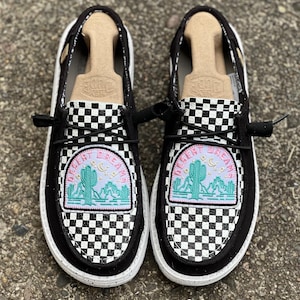 May include: A pair of black and white checkered slip-on shoes with a white sole. Each shoe features a patch with a desert scene and the words "Desert Dreams". The shoes have black laces and a canvas upper.