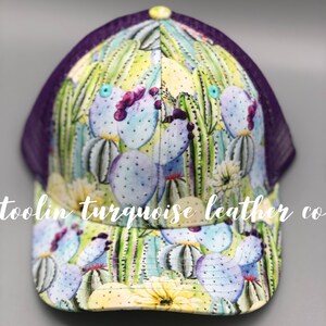 May include: A trucker hat featuring a vibrant cactus print with purple mesh backing. The hat showcases a colorful design of cacti in shades of green, blue, and purple, with a yellow brim and a button on top. The text "toolin turquoise leather co" is printed on the hat.