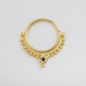 A gold-colored nose ring with a blue gemstone is shown against a plain white background.