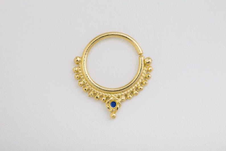 A gold-colored nose ring with a blue gemstone is shown against a plain white background.
