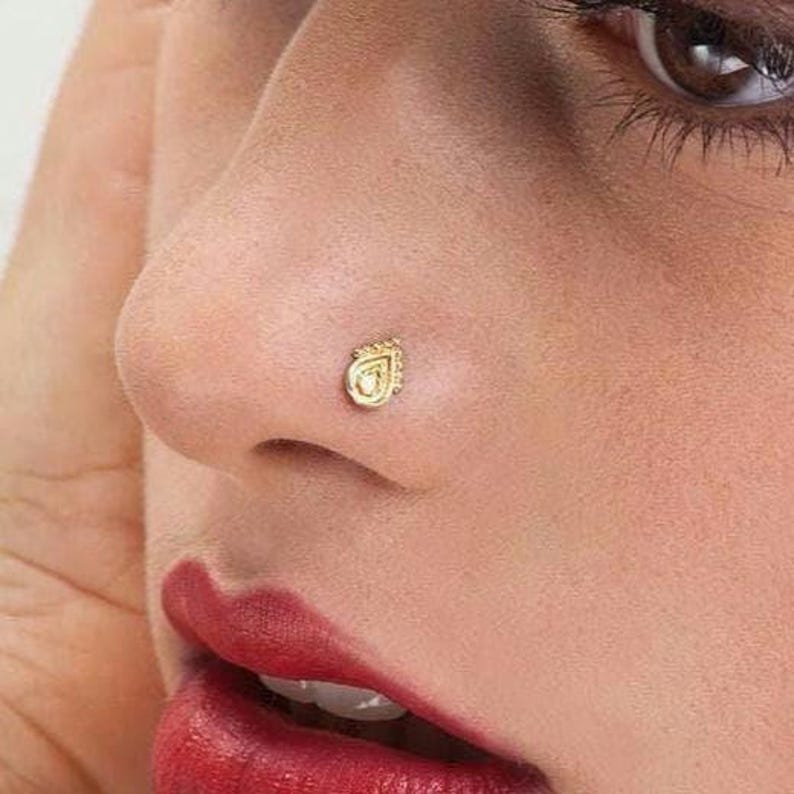 Dainty Gold Nose Stud: A close-up of a woman's face, with her eyes closed and her hands covering her ears. She has red lipstick and a nose stud.