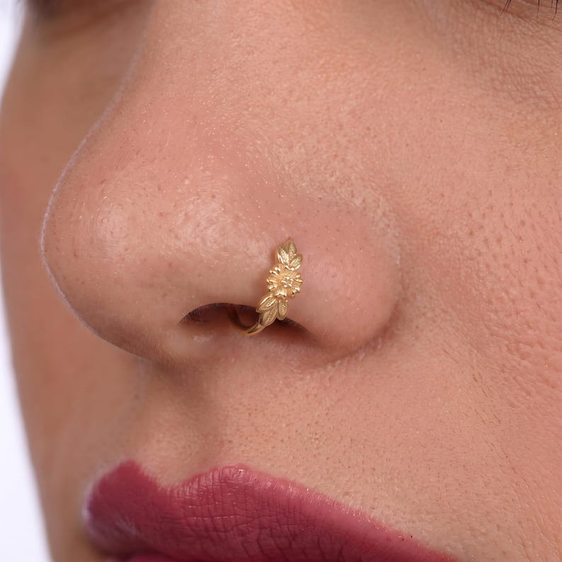 A close-up of a person's nose with a gold-colored nose ring featuring a leaf design.