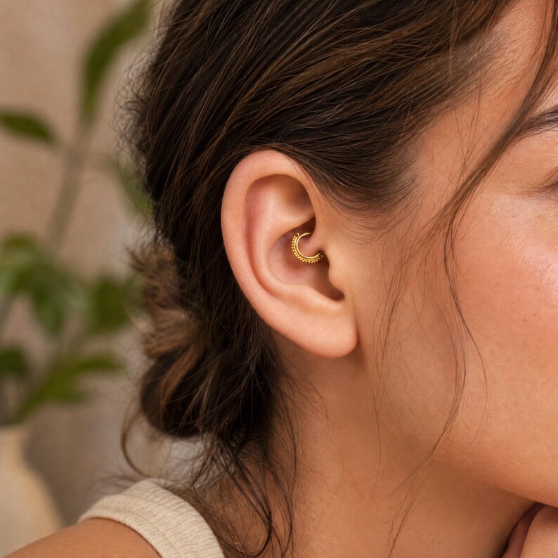 A close-up of a woman's ear with a gold earring in it, wearing a beige top.
