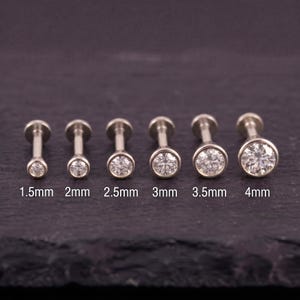 Può includere: Un set di sei borchie labret color argento con pietre rotonde trasparenti in incastonatura a lunetta, disposte in ordine di grandezza crescente da 1,5 mm a 4 mm. Ogni borchia presenta un perno piatto e una pietra circolare, con le misure indicate.