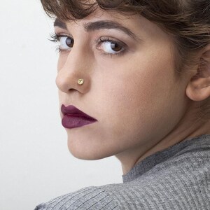 A close-up portrait of a woman with dark hair and a nose piercing, wearing a gray sweater.