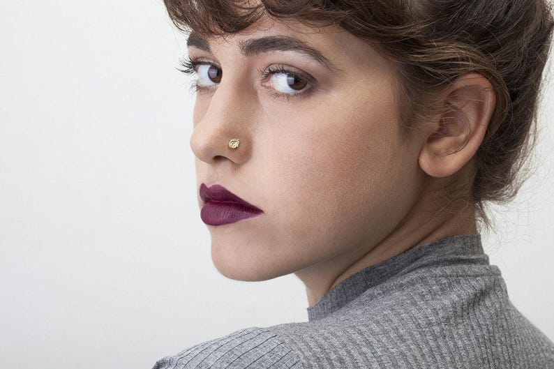 A close-up portrait of a woman with dark hair and a nose piercing, wearing a gray sweater.