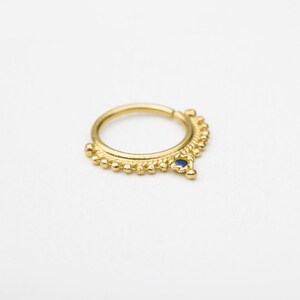 A gold-colored metal nose ring with a blue gemstone is shown against a white background.