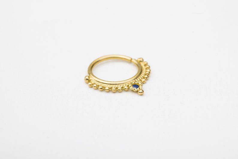 A gold-colored metal nose ring with a blue gemstone is shown against a white background.