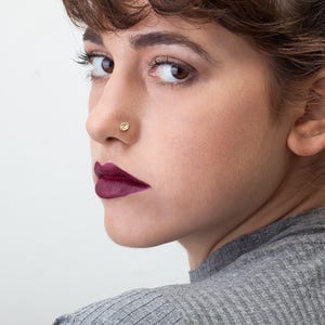 A close-up portrait of a woman with brown hair and a nose piercing, wearing a gray sweater and looking off to the side with a serious expression.