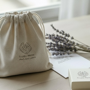 A beige drawstring bag with the logo "Studio Meme" printed on it, along with a bouquet of lavender flowers and a white card or box with the same logo.