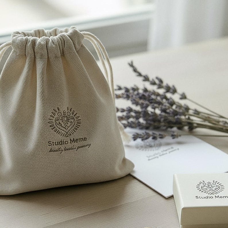 A beige drawstring bag with the logo "Studio Meme" printed on it, along with a bouquet of lavender flowers and a white card or box with the same logo.