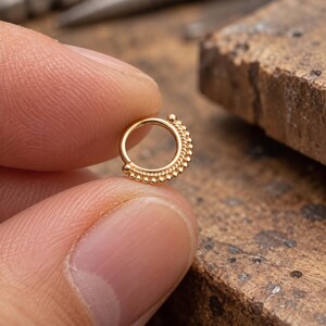 A close-up image of a person's finger holding a small, ornate gold-colored piercing.