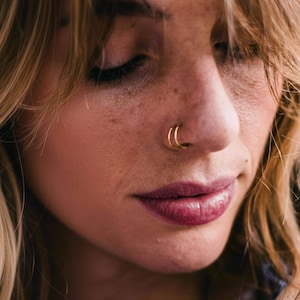 May include: Close-up of a person's face featuring a double gold nose ring. The person has fair skin, freckles, and closed eyes. Their lips are a deep shade of red, and their hair is blonde and wavy.