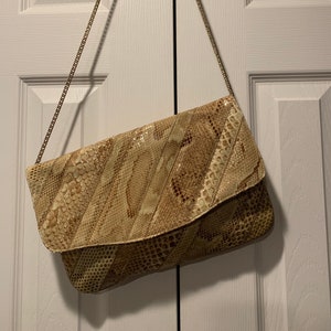 May include: A beige snakeskin clutch bag with a gold chain strap. The bag has a flap closure and a textured snakeskin pattern.